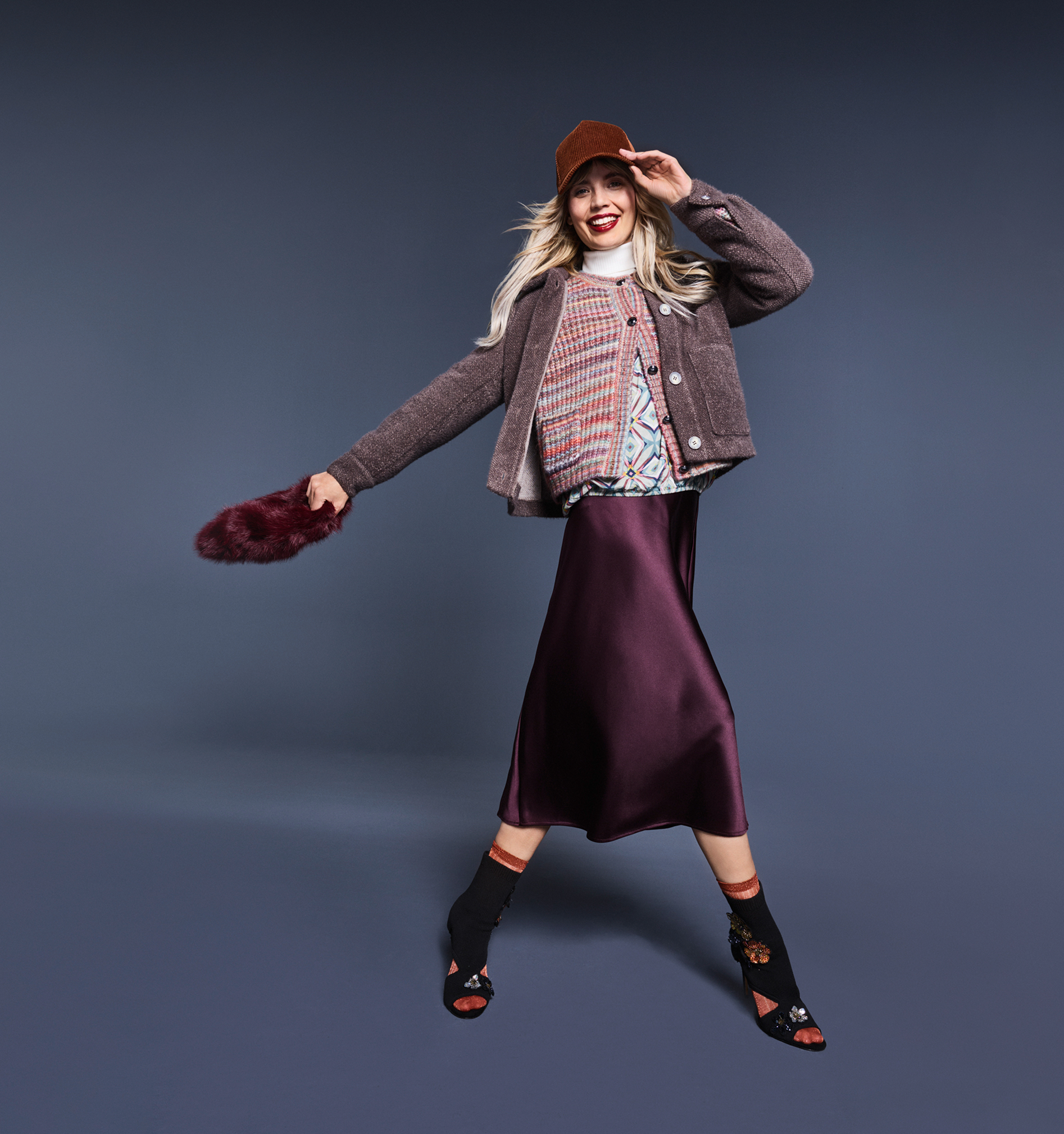 Eine Frau steht in einem Studio und hat einen Satin Rock mit einer Strickhacke und einer Jacke an.