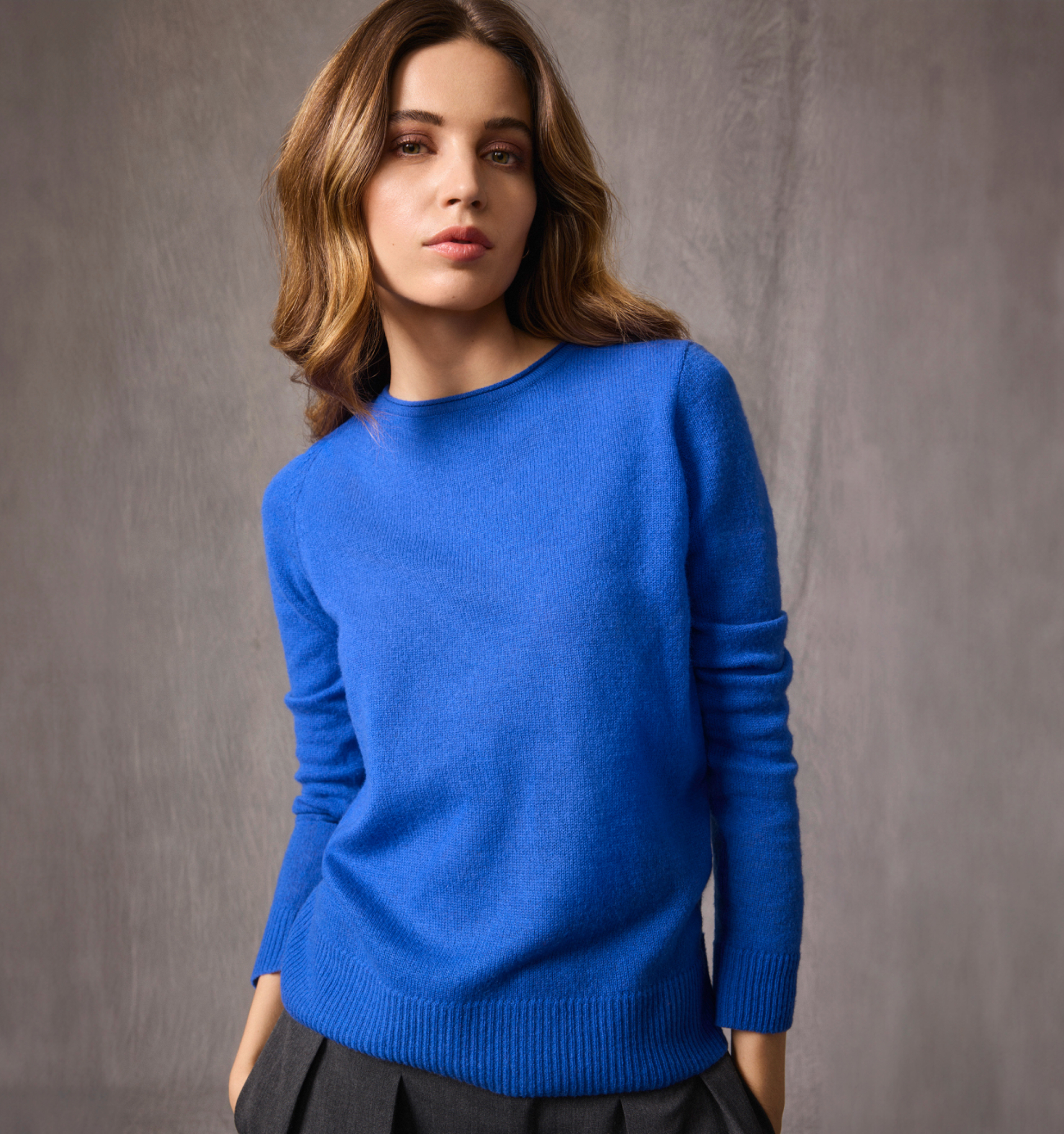 Eine Frau steht mit einem blauen Kaschmirpullover vor einer grauen Wand.